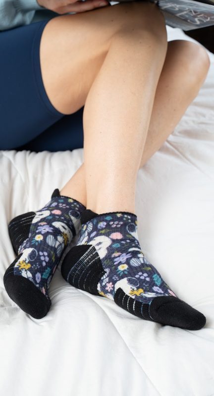 Legs crossed on a white bed in navy floral ankle compression socks with black sole bands